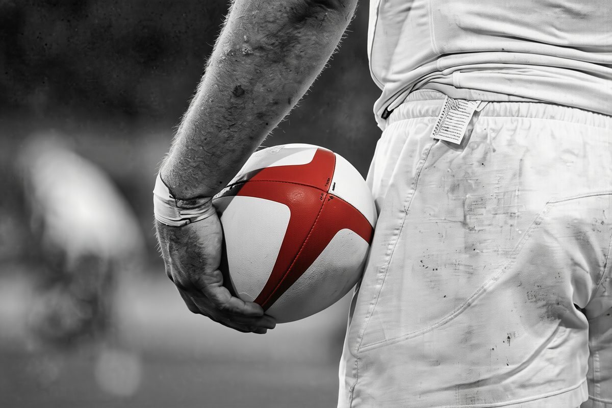 A player in white shorts holding a rugby ball against his hip
